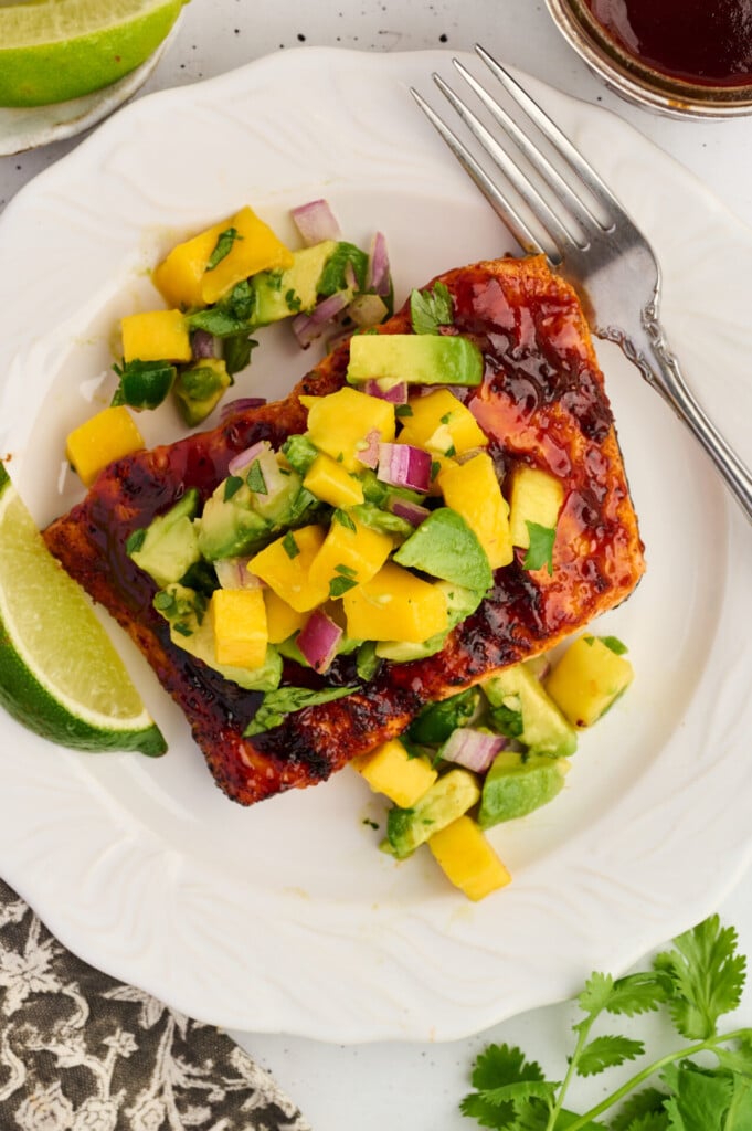 A white plate with grilled barbecue salmon steaks topped with a vibrant avocado-mango salsa of diced mango, avocado, red onion, and cilantro, garnished with a lime wedge. A fork rests on the plate.