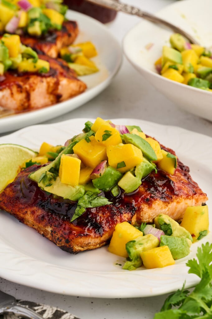 Grilled Barbecue Salmon Steaks with Avocado-Mango Salsa feature a perfectly cooked salmon fillet topped with fresh mango avocado salsa, garnished with lime, and served on a white plate with extra salsa and salmon steaks in the background.