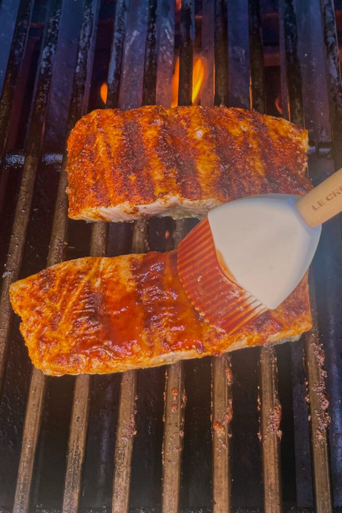 Two pieces of seasoned salmon are grilling over open flames, while a silicone brush is spreading sauce over one filet—perfect for making Grilled Barbecue Salmon Steaks with Avocado-Mango Salsa.