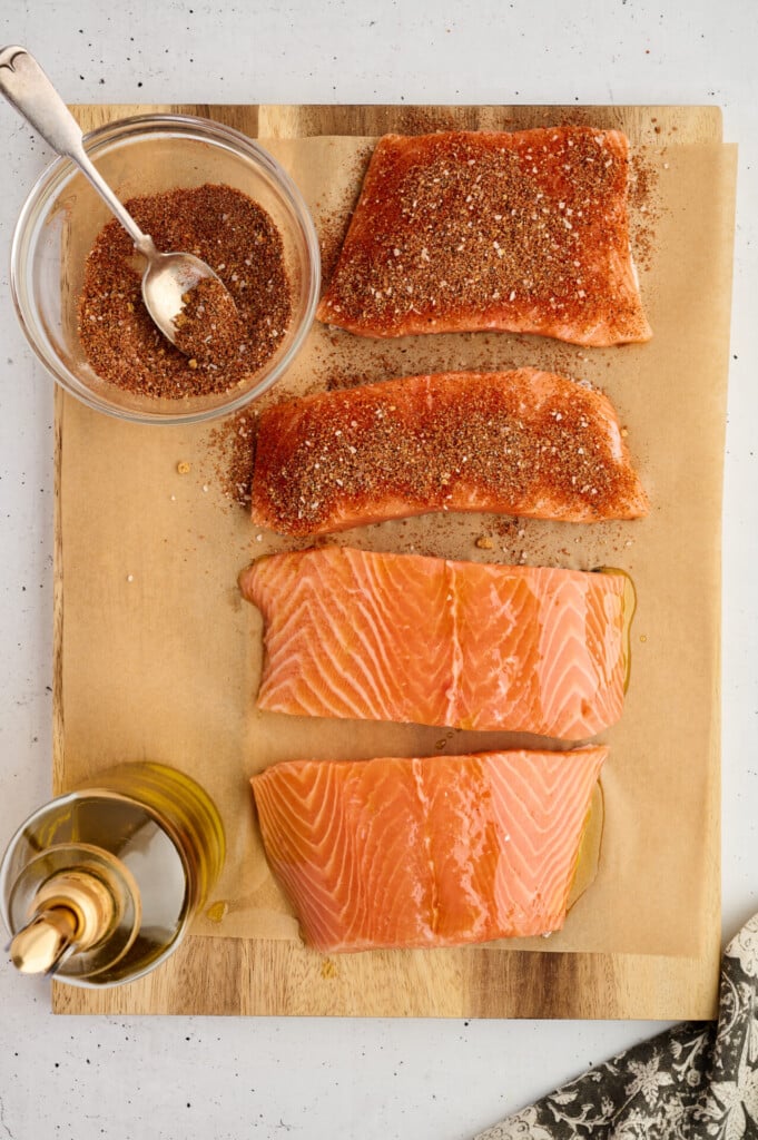 Two raw salmon fillets and two seasoned grilled barbecue salmon steaks with avocado-mango salsa rest on parchment paper, accompanied by a bowl of spice mix and a spoon above them, with a bottle of oil to the left.