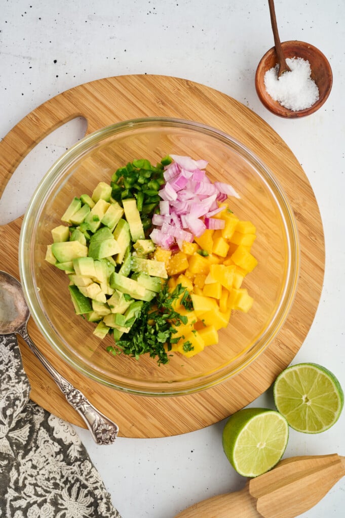 A glass bowl with diced avocado, mango, red onion, jalapeño, and cilantro on a round wooden board—perfect as a vibrant topping for Grilled Barbecue Salmon Steaks with Avocado-Mango Salsa—with salt, lime halves, and a spoon nearby.
