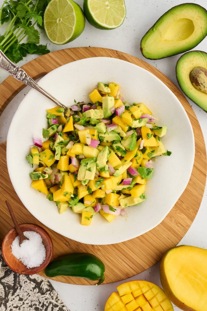 A white bowl of mango avocado salsa with red onion and cilantro sits on a wooden board, ready to complement any summer meal. Surrounding it are lime halves, avocado, fresh cilantro, a jalapeño, a mango half, and a small bowl of salt—the perfect fresh ingredients for topping Grilled Barbecue Salmon Steaks with Avocado-Mango Salsa.