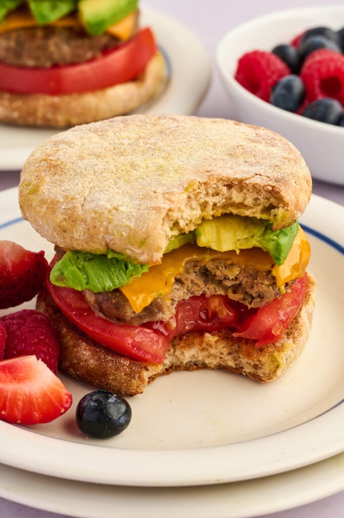 A breakfast sandwich with a bite taken out, showing layers of egg, cheddar, avocado, and tomato on an English muffin with savory Apple Breakfast Sausage Patties. Fresh berries are on the plate, with more berries in a bowl in the background.