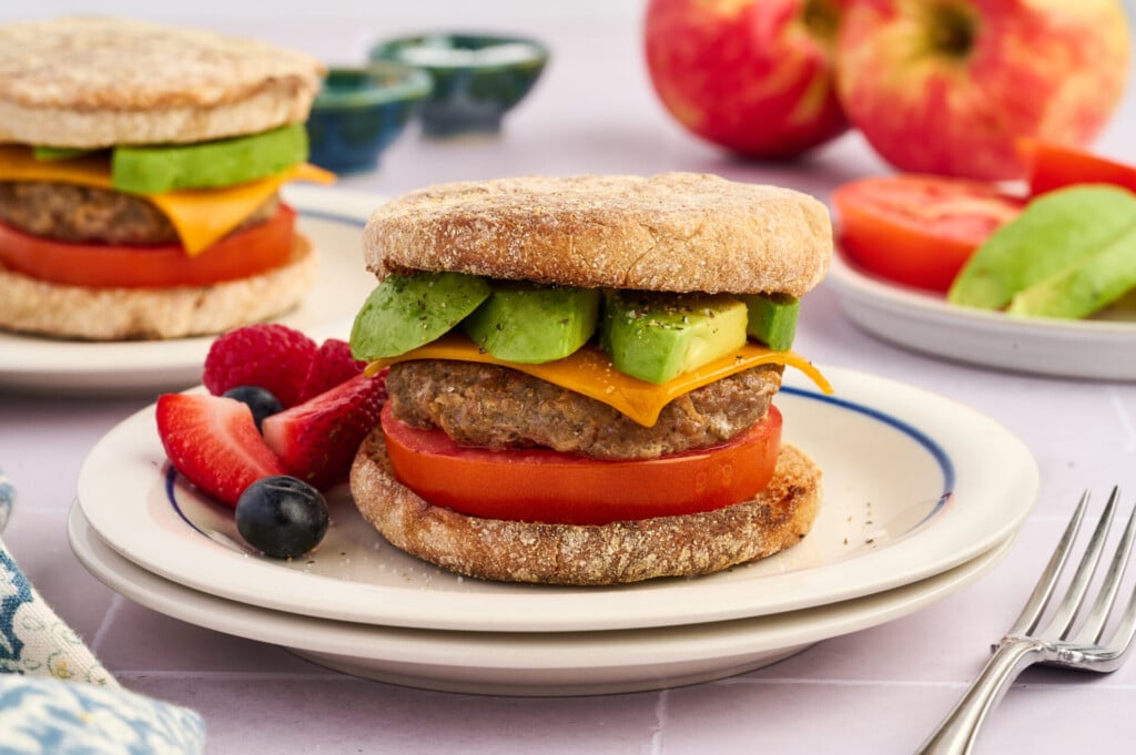An English muffin sandwich with an Apple Breakfast Sausage Patty, cheddar cheese, avocado slices, and tomato, served on a plate with fresh berries. Another sandwich and fruit are in the background.