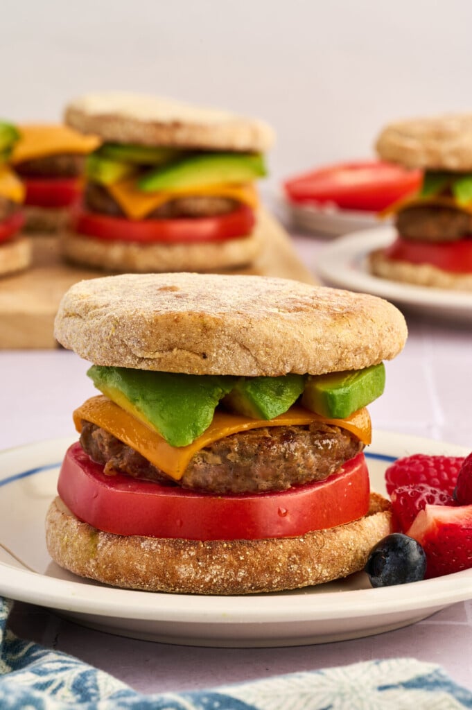 A breakfast sandwich featuring Apple Breakfast Sausage Patties, cheddar cheese, avocado, and tomato on an English muffin, served on a plate with fresh berries. Other similar sandwiches are in the background.