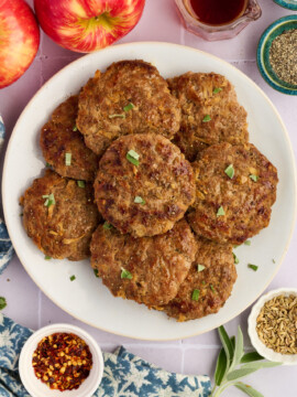 A plate of browned Apple Breakfast Sausage Patties garnished with herbs, surrounded by apples, a small bowl of red pepper flakes, fennel seeds, black pepper, and a glass jar of syrup on a light surface.