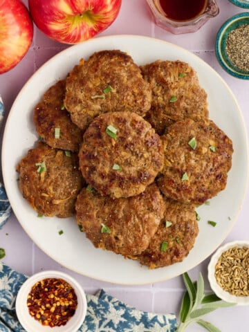 A plate of browned Apple Breakfast Sausage Patties garnished with herbs, surrounded by apples, a small bowl of red pepper flakes, fennel seeds, black pepper, and a glass jar of syrup on a light surface.