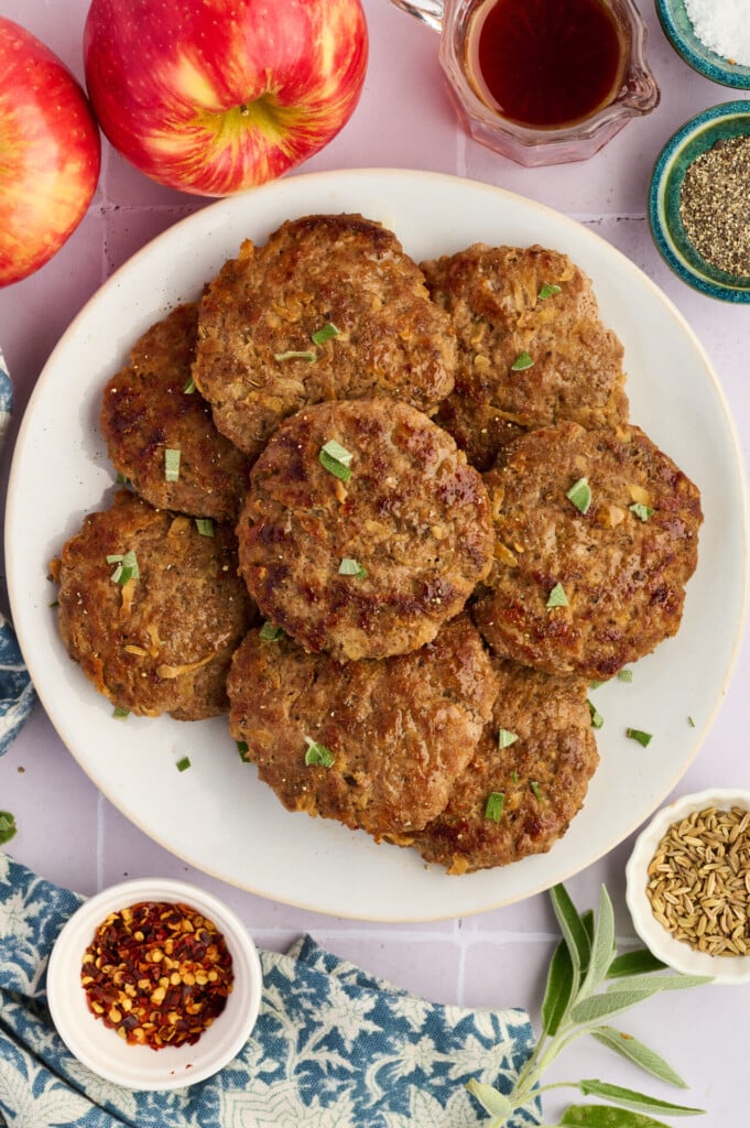 A plate of browned Apple Breakfast Sausage Patties garnished with herbs, surrounded by apples, a small bowl of red pepper flakes, fennel seeds, black pepper, and a glass jar of syrup on a light surface.
