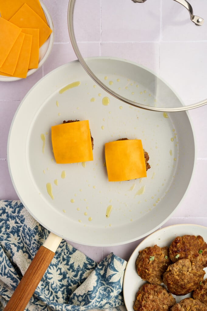 A frying pan with two Apple Breakfast Sausage Patties topped with cheddar cheese slices, under a glass lid. Nearby are more patties on a plate, extra cheese slices, and a blue patterned towel.