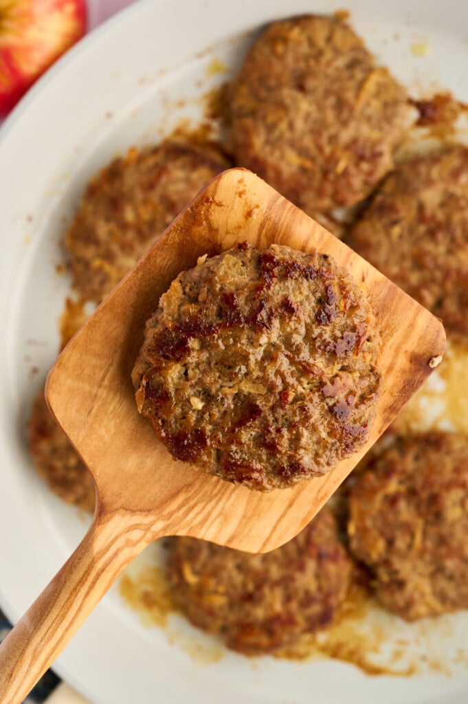 A wooden spatula holds a cooked Apple Breakfast Sausage Patty above a plate filled with more patties. The patties appear browned and have a slightly rough texture.