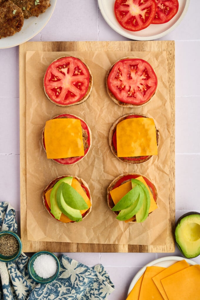 Six open-faced burgers on parchment: top row plain with tomato slices, middle row with tomato and cheddar cheese, bottom row with cheddar cheese, tomato, and avocado slices. Surrounding items include Apple Breakfast Sausage Patties, fresh tomato, creamy avocado, condiments, and cheese.