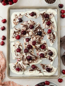 A baking sheet with pieces of Frozen Chocolate Cherry Yogurt Bark, featuring swirled chocolate and topped with fresh cherries, surrounded by bowls of cherries and chocolate chips on a light surface.