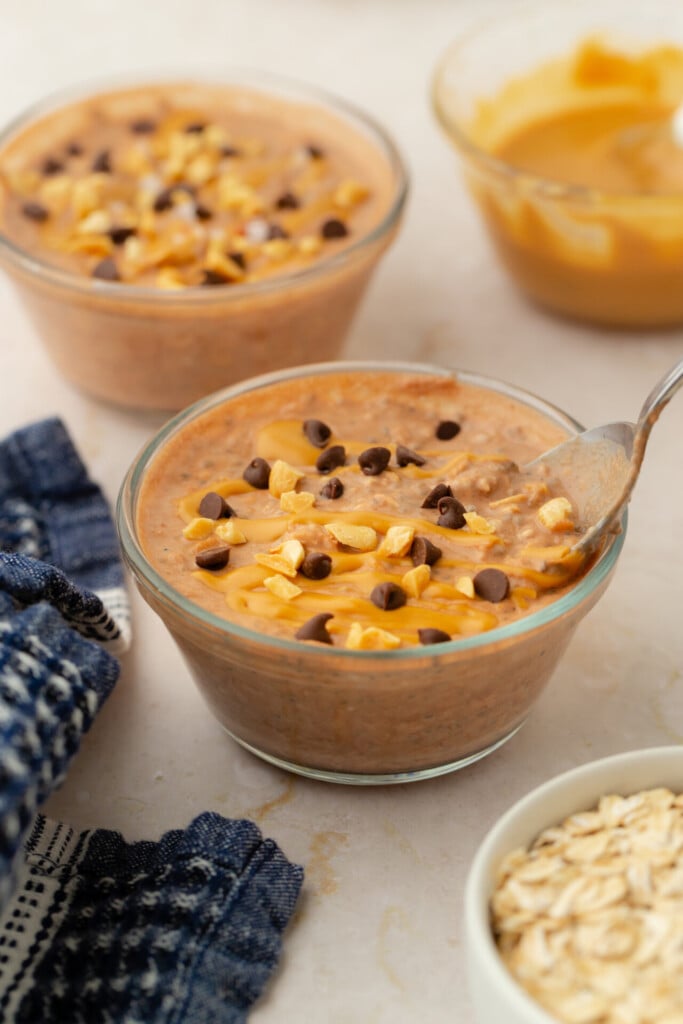 A glass bowl of Chocolate Peanut Butter Overnight Oats topped with chocolate chips and chopped peanuts, with a spoon inside. Other bowls of these rich, creamy oats are nearby on a light surface, each brimming with similar delicious ingredients.