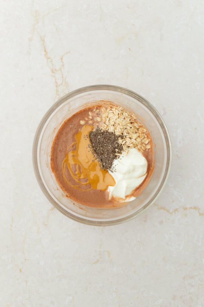 A glass bowl with cocoa liquid, oats, chia seeds, yogurt, and honey sits on a light marble surface—the perfect base for Chocolate Peanut Butter Overnight Oats. The ingredients are unmixed and grouped separately in the bowl.