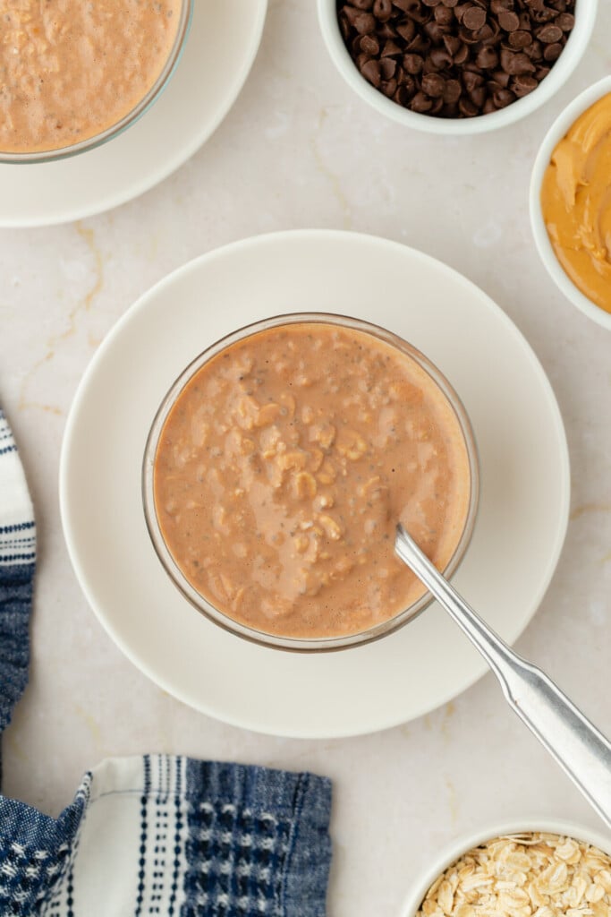 A bowl of chocolate oatmeal with a spoon, surrounded by small bowls of chocolate chips, oats, and peanut butter, evokes the indulgence of Chocolate Peanut Butter Overnight Oats, all set off by a blue plaid napkin.