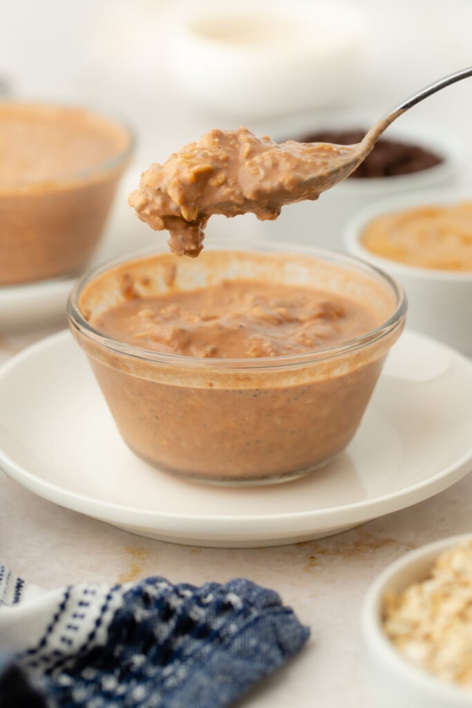 A spoon holds a scoop of creamy, brownish Chocolate Peanut Butter Overnight Oats above a small glass bowl filled with the same mixture. Several other bowls with different contents are in the background.