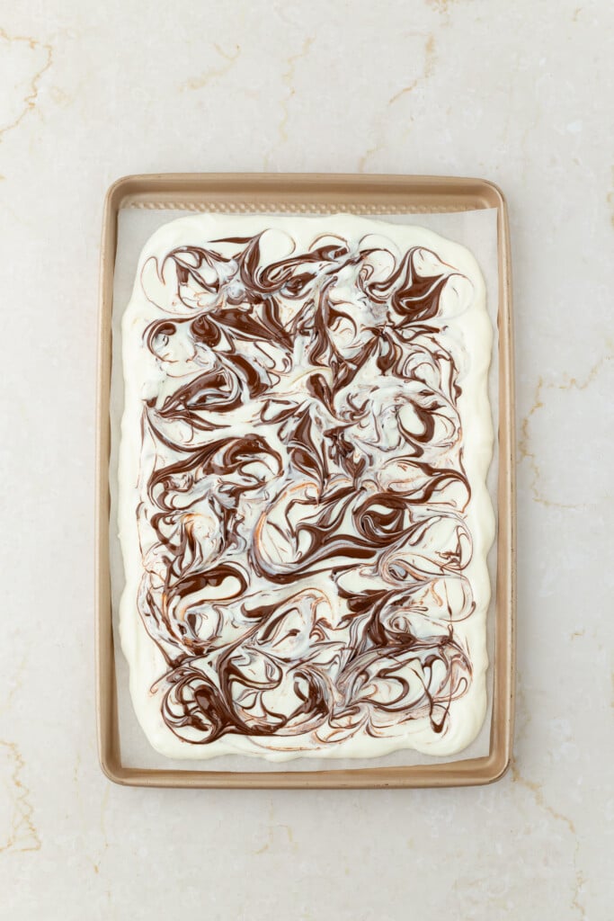 A rectangular baking sheet with a layer of white batter swirled with chocolate, creating a marbled pattern reminiscent of Frozen Chocolate Cherry Yogurt Bark, sits on a light-colored marble surface.