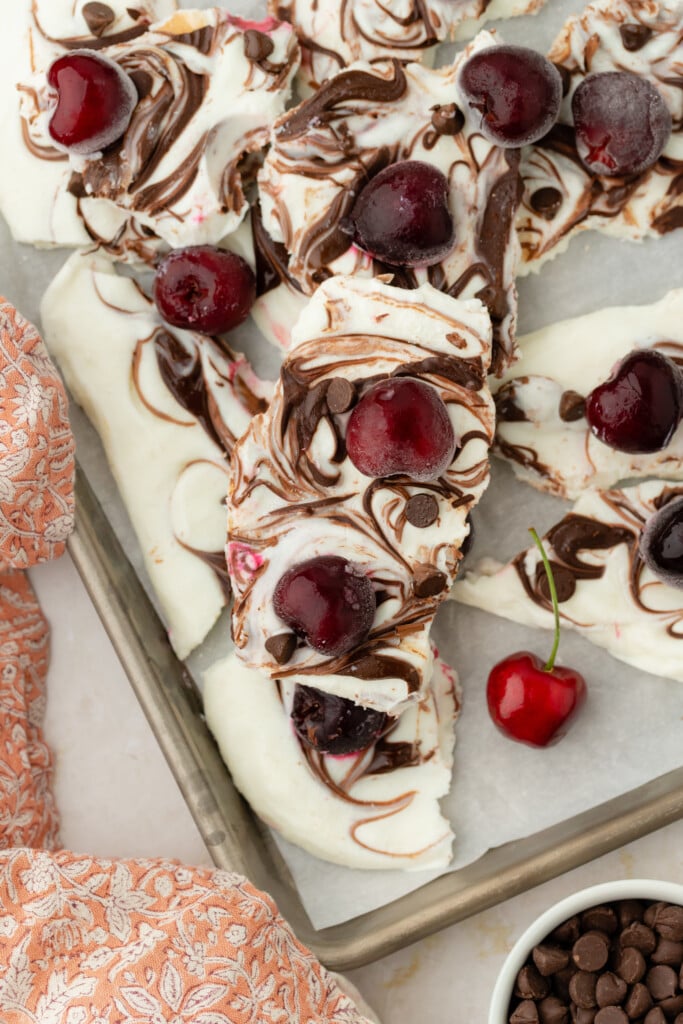Rectangular pieces of marbled Frozen Chocolate Cherry Yogurt Bark topped with whole cherries and chocolate chips rest on a parchment-lined baking sheet, with a red patterned cloth and a bowl of chocolate chips nearby.