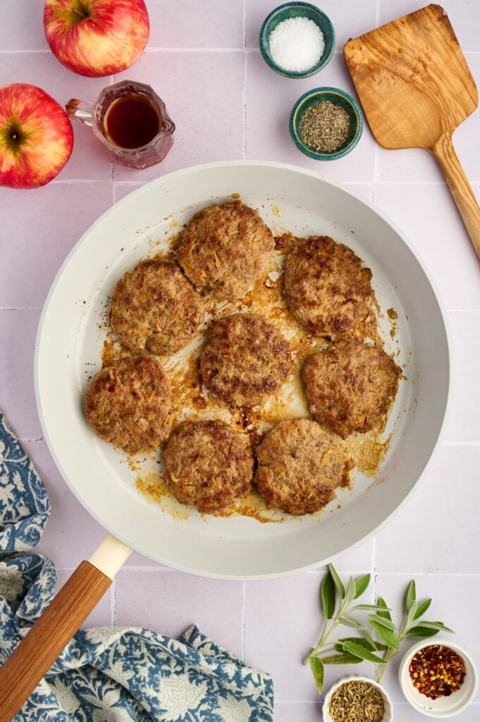 Seven browned Apple Breakfast Sausage Patties sizzle in a white skillet, surrounded by apples, seasonings, a wooden spatula, and a blue patterned cloth on a light tiled surface.