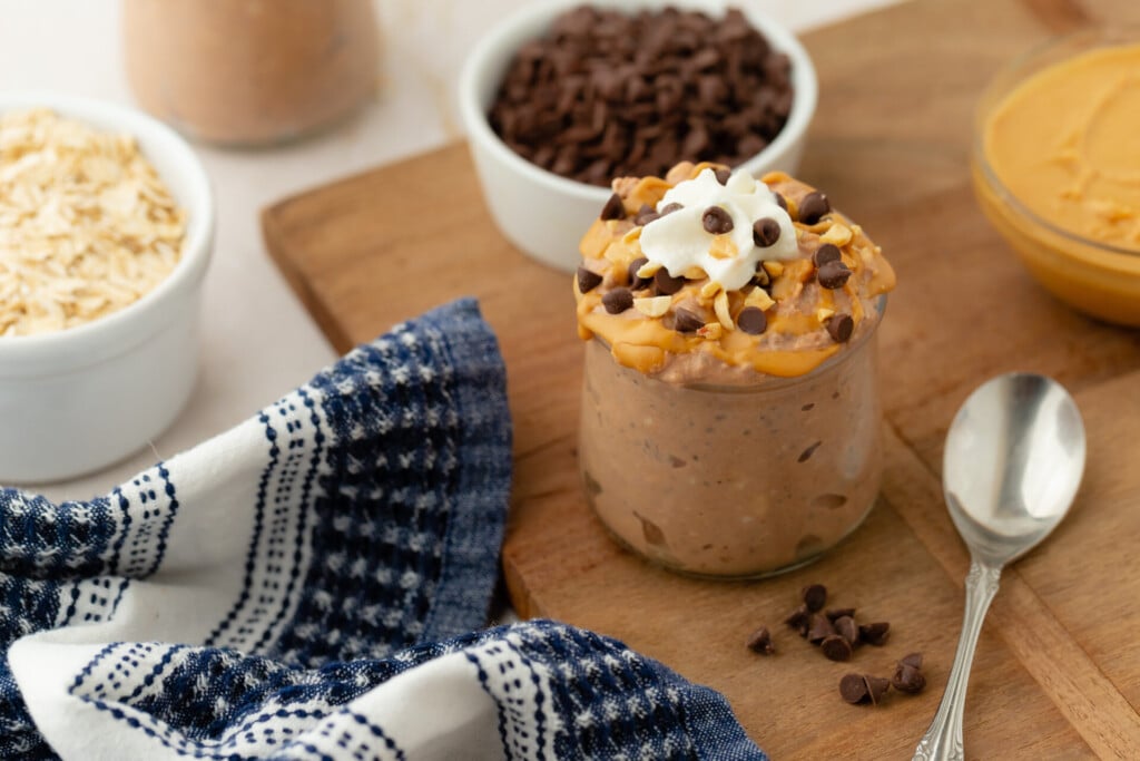 A jar of Chocolate Peanut Butter Overnight Oats topped with whipped cream and chocolate chips sits on a wooden board next to a spoon, a blue patterned napkin, and small bowls of oats, chocolate chips, and peanut butter.