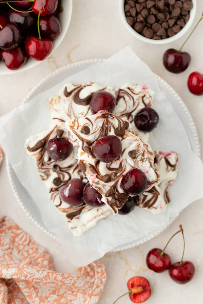 A plate of chocolate-swirled cherry bars inspired by Frozen Chocolate Cherry Yogurt Bark, topped with fresh cherries and set on parchment paper. Bowls of cherries and chocolate chips surround the plate, with a peach-colored patterned napkin nearby.