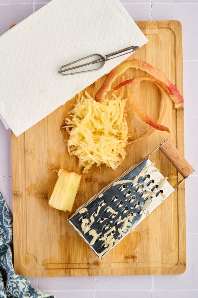 A wooden cutting board with grated apple, a grater, apple peel, a core, a peeler, and a paper towel on top hints at prep for Apple Breakfast Sausage Patties, with part of a blue-patterned cloth visible in the corner.