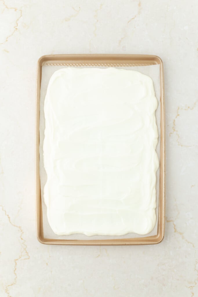 A baking sheet lined with parchment paper holds an even layer of melted white chocolate, perfect for making Frozen Chocolate Cherry Yogurt Bark. The smooth surface rests on a light-colored, marbled countertop.