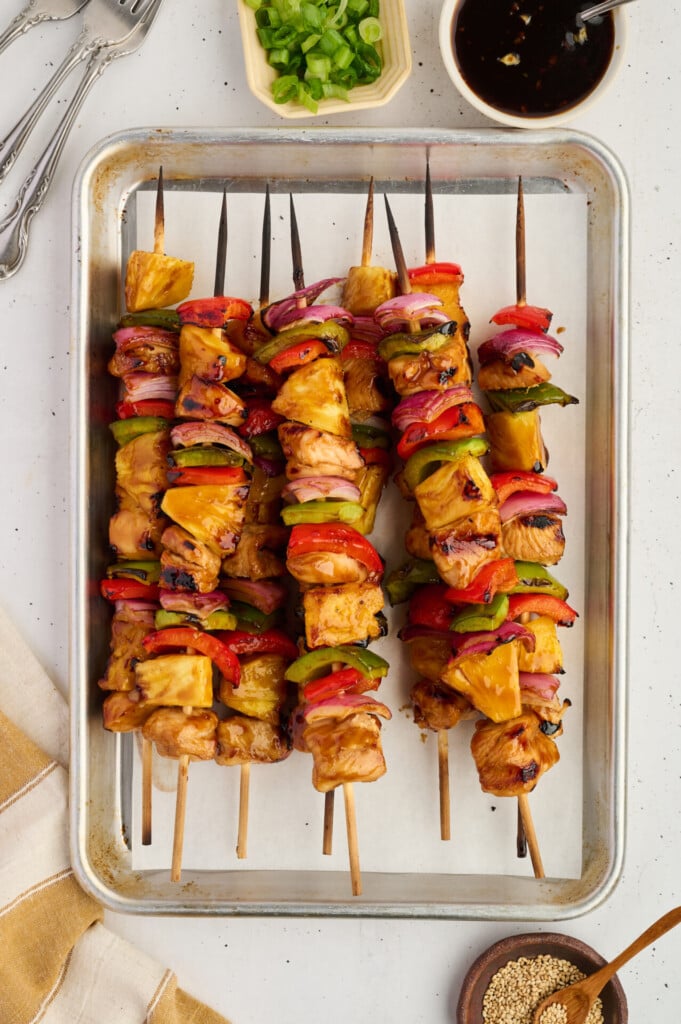 Five skewers with Healthy High Protein Grilled Teriyaki Chicken & Pineapple, red onion, and bell peppers rest on a parchment-lined baking tray. Garnishes and dipping sauce are nearby on the table.