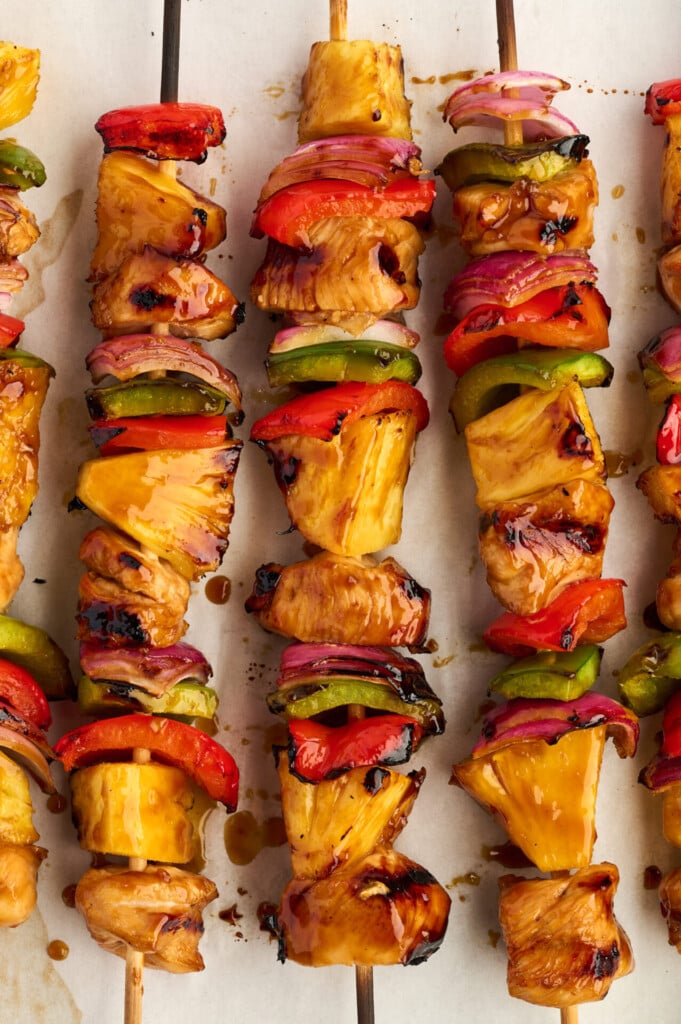 Healthy High Protein Grilled Teriyaki Chicken & Pineapple Skewers feature glazed chicken, pineapple chunks, red onion, red bell pepper, and green bell pepper—all perfectly grilled and served on parchment paper.