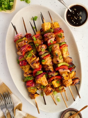 Five skewers of healthy high protein Grilled Teriyaki Chicken & Pineapple Skewers, featuring juicy grilled chicken, sweet pineapple, red onion, and bell peppers on an oval plate, garnished with green onions. Dipping sauce, sesame seeds, forks, and a napkin are nearby on a white surface.