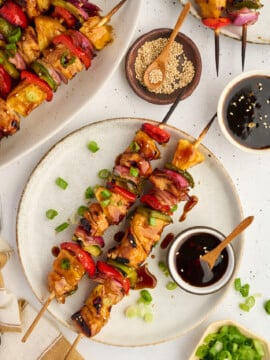 Two plates with Healthy High Protein Grilled Teriyaki Chicken & Pineapple Skewers and assorted vegetables, garnished with green onions. Dipping sauce, sesame seeds, and chopped green onions are placed nearby on a white table.