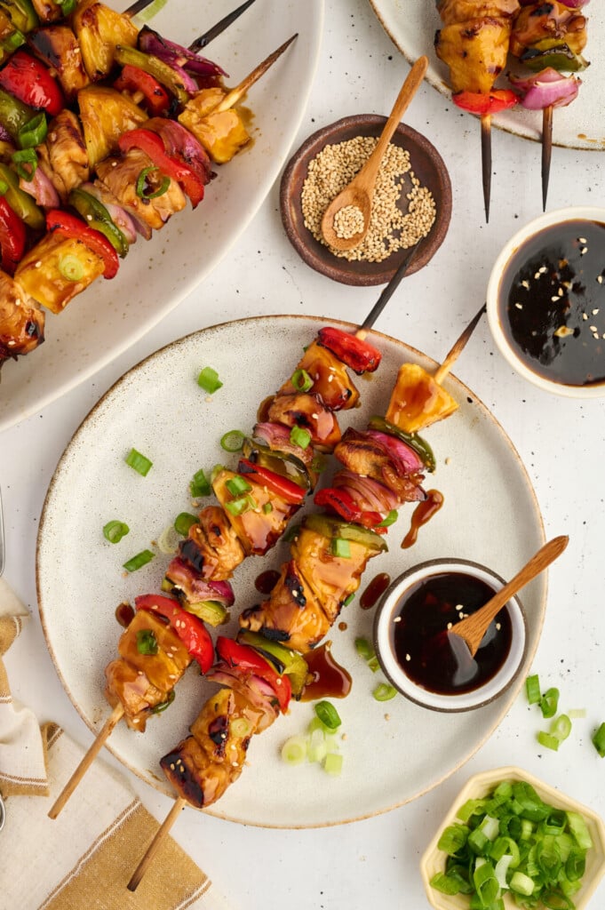 Two plates with Healthy High Protein Grilled Teriyaki Chicken & Pineapple Skewers and assorted vegetables, garnished with green onions. Dipping sauce, sesame seeds, and chopped green onions are placed nearby on a white table.