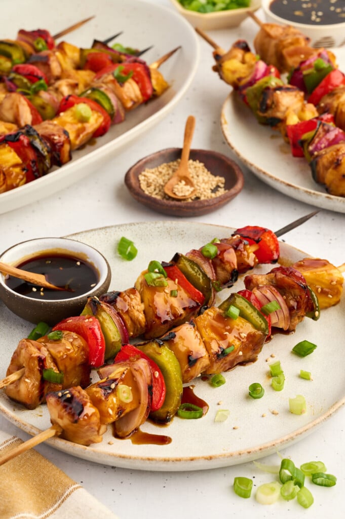 Healthy High Protein Grilled Teriyaki Chicken & Pineapple Skewers are served on plates, glazed with savory sauce and paired with a bowl of dipping sauce. Finished with a sprinkle of sesame seeds and chopped green onions on the side, these skewers offer a delicious blend of chicken, vegetables, and juicy pineapple for a nutritious meal.