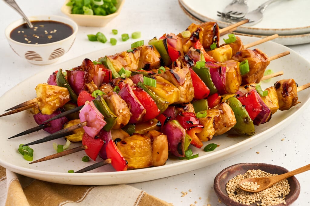 Grilled vegetable and pineapple skewers with sauce, garnished with chopped green onions and sesame seeds, served on a white platter alongside Healthy High Protein Grilled Teriyaki Chicken & Pineapple Skewers, with a bowl of dipping sauce and plates in the background.