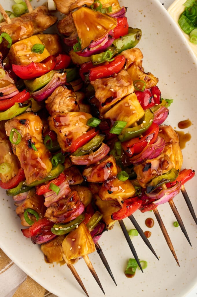 Enjoy our Healthy High Protein Grilled Teriyaki Chicken & Pineapple Skewers—three grilled skewers with chunks of chicken, pineapple, red and green bell peppers, and red onion, topped with chopped green onions and a drizzle of teriyaki sauce, served on a white plate.