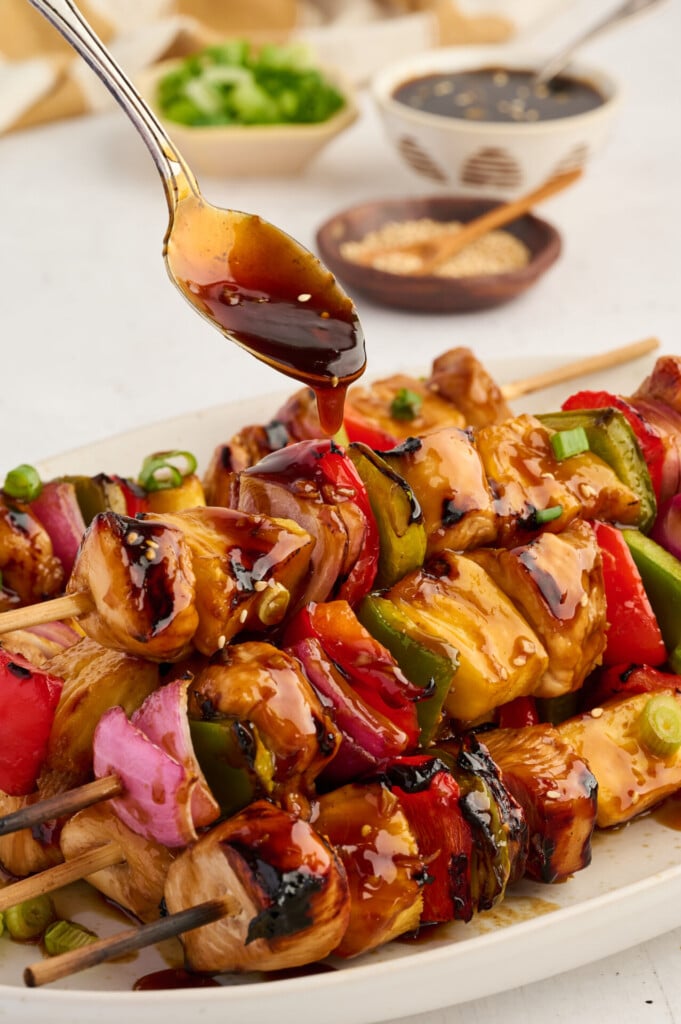 Chicken and vegetable skewers on a plate are being drizzled with a dark sauce from a spoon. These Healthy High Protein Grilled Teriyaki Chicken & Pineapple Skewers include pieces of chicken, red onion, green and red bell peppers, and juicy pineapple. Bowls of sauce and toppings are in the background.