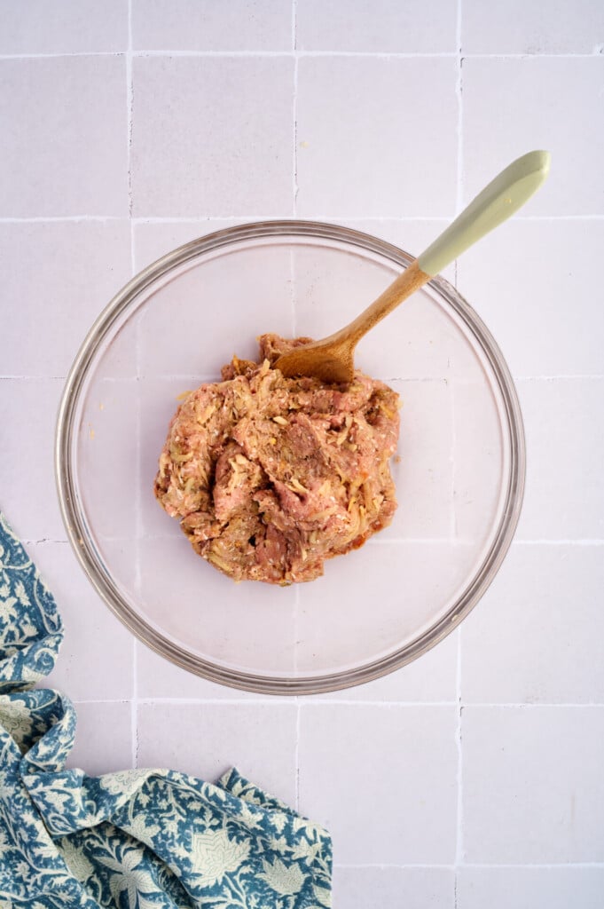 A glass bowl with raw ground meat mixture for Apple Breakfast Sausage Patties and a wooden spoon, sitting on a light tiled surface next to a blue and white patterned cloth.
