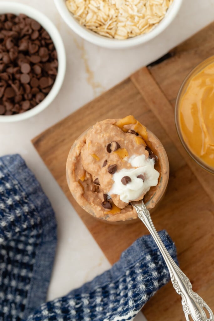 A small bowl of Chocolate Peanut Butter Overnight Oats, topped with whipped cream and chocolate chips, sits next to bowls of oats and chocolate chips on a wooden board, with a blue cloth nearby.