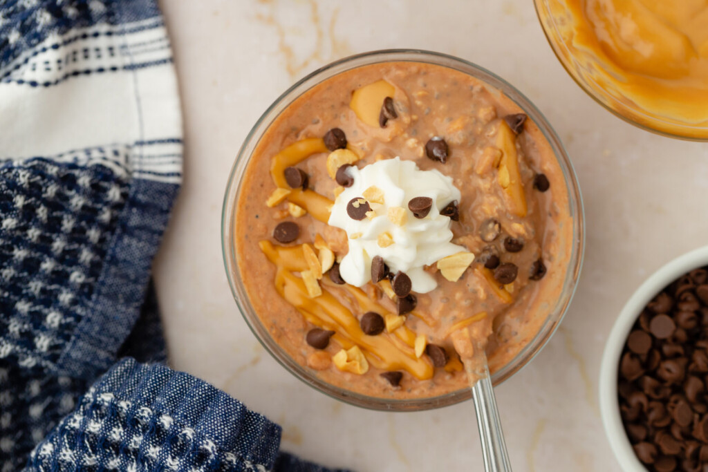 A decadent bowl of Chocolate Peanut Butter Overnight Oats topped with whipped cream, chocolate chips, a luscious peanut butter drizzle, and chopped nuts sits beside a blue checkered cloth and a small bowl of chocolate chips.
