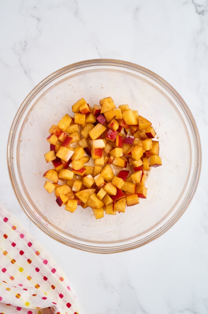 A glass bowl filled with diced apples coated in spices sits on a white marble surface next to a colorful dotted kitchen towel, evoking the cozy flavors of an Easy 5-Minute Peach Cobbler.