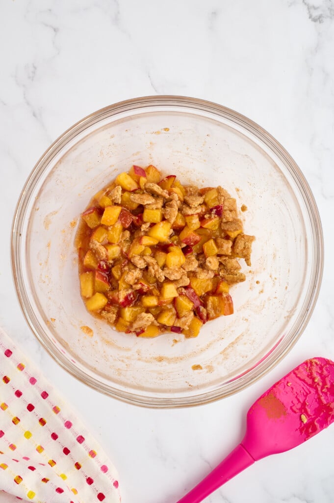 A glass bowl with chopped peaches mixed with cinnamon and sugar sits on a marble surface, ready for your Easy 5-Minute Peach Cobbler recipe. A pink spatula and a polka-dotted cloth are nearby.