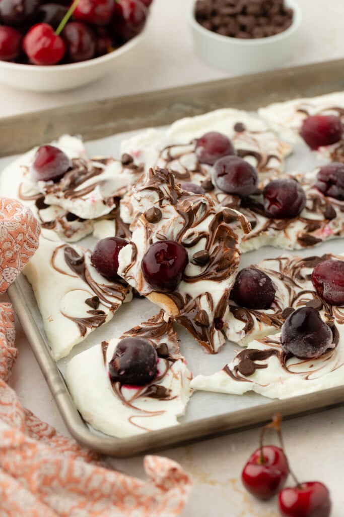 A baking sheet with pieces of swirled Frozen Chocolate Cherry Yogurt Bark, topped with whole cherries and chocolate chips. A bowl of cherries and a bowl of chocolate chips are in the background, next to a patterned cloth.