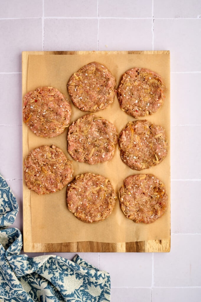Seven raw burger patties, including flavorful Apple Breakfast Sausage Patties, rest on a parchment-lined wooden board placed on a light tile surface, with a blue and white patterned cloth to the side.