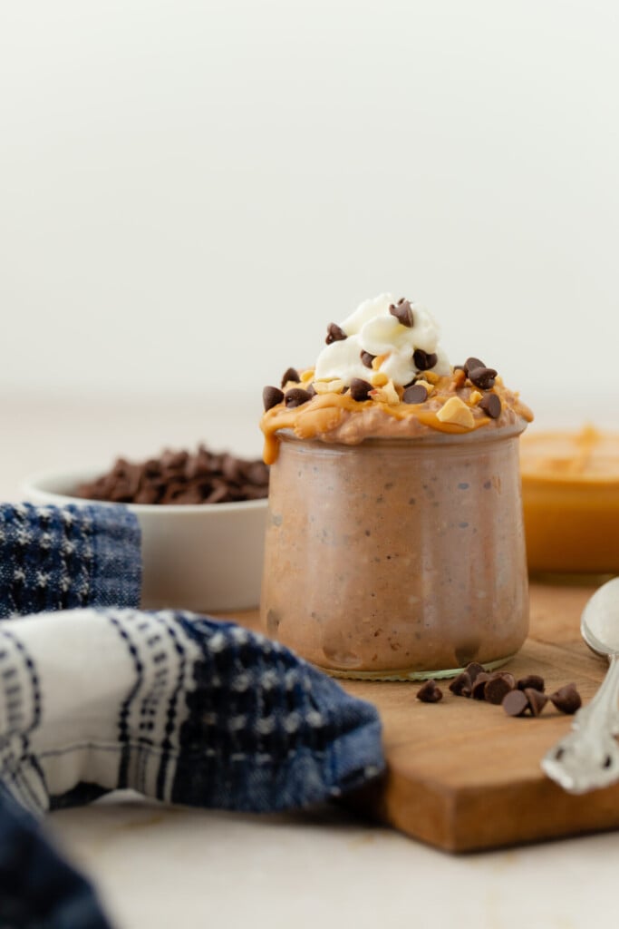 A glass jar filled with Chocolate Peanut Butter Overnight Oats, topped with whipped cream, chocolate chips, and peanut butter. A spoon, blue plaid napkin, and bowls of chocolate chips and peanut butter are nearby.
