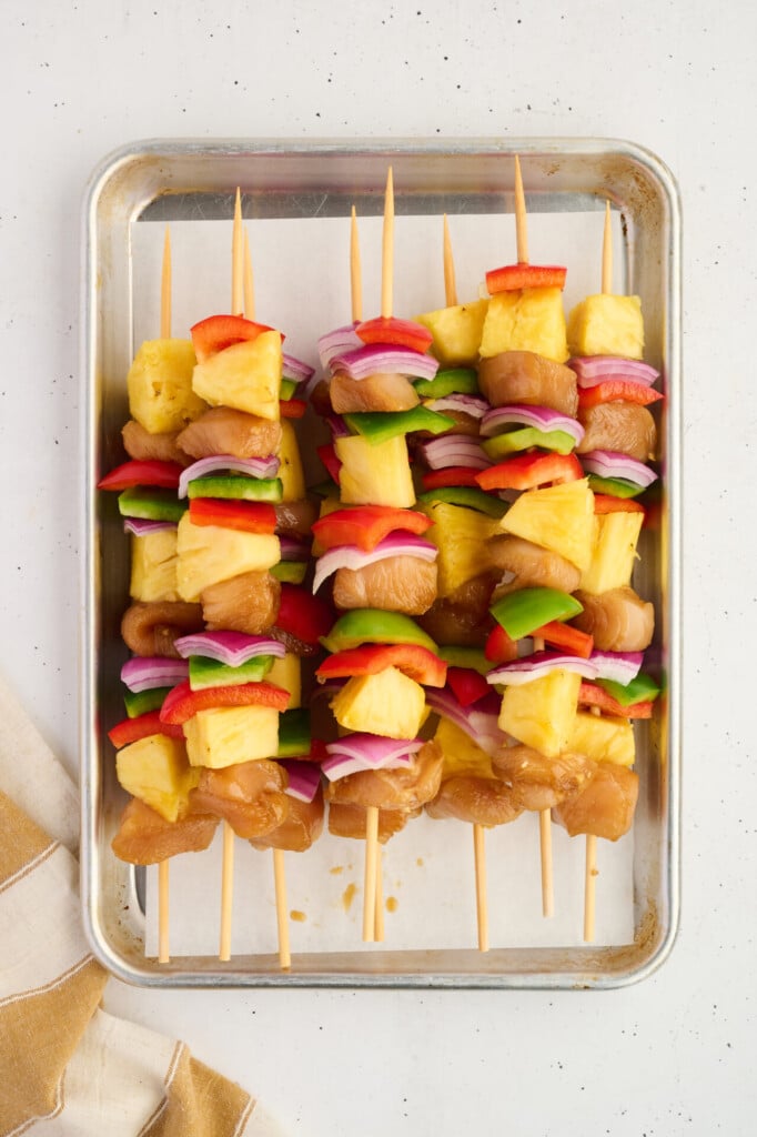 Raw chicken, pineapple chunks, red onion, green bell pepper, and red bell pepper pieces are threaded onto wooden skewers and arranged on a metal baking sheet, ready for grilling or baking into Healthy High Protein Grilled Teriyaki Chicken & Pineapple Skewers.