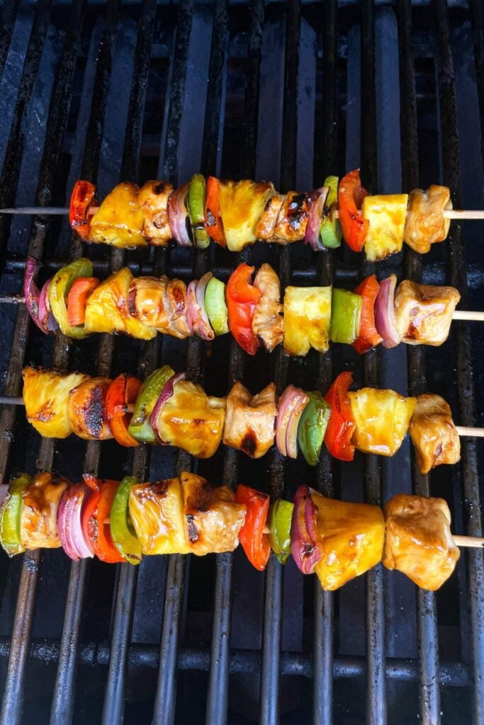 Three skewers with Healthy High Protein Grilled Teriyaki Chicken & Pineapple, red onion, bell pepper, and mushrooms on a barbecue grill. The colorful skewers are perfectly cooked and sit enticingly over the grill grates.