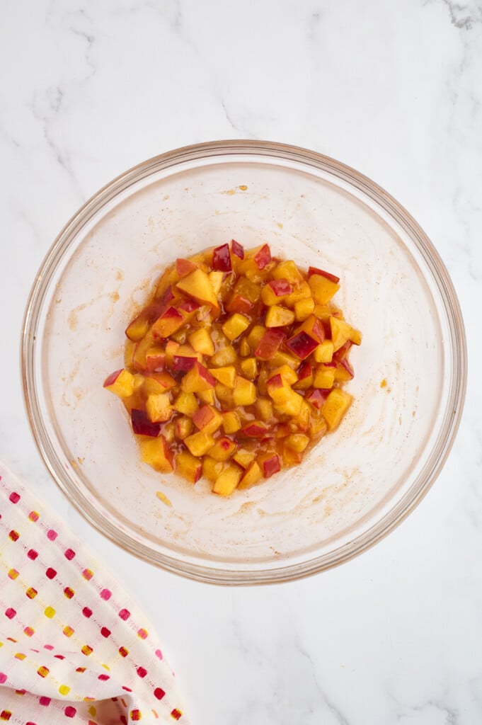 A glass bowl filled with diced apples mixed with cinnamon and sugar sits on a white marble surface next to a colorful dotted cloth, evoking the comforting flavors of an Easy 5-Minute Peach Cobbler.