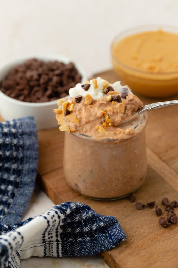 A glass jar filled with creamy Chocolate Peanut Butter Overnight Oats, topped with whipped cream, nuts, and chocolate chips. Bowls of chocolate chips and peanut butter are in the background, along with a blue patterned cloth.