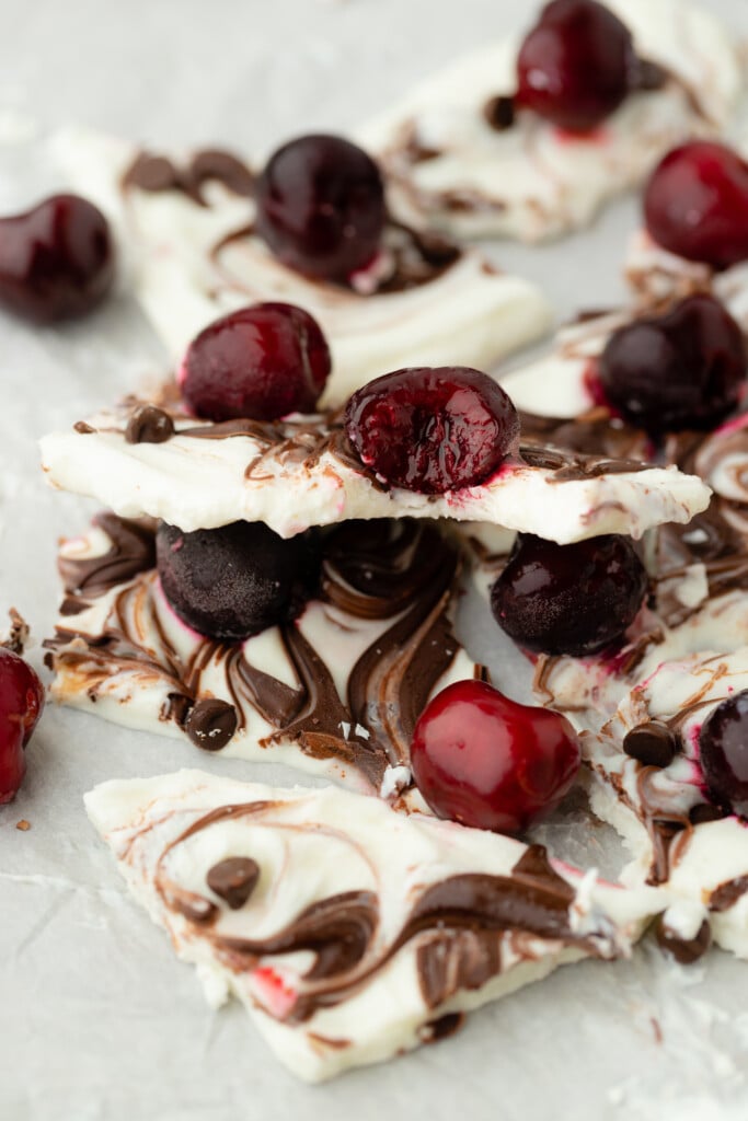 Broken pieces of swirled white and dark Frozen Chocolate Cherry Yogurt Bark, topped with whole cherries and scattered on a light surface.