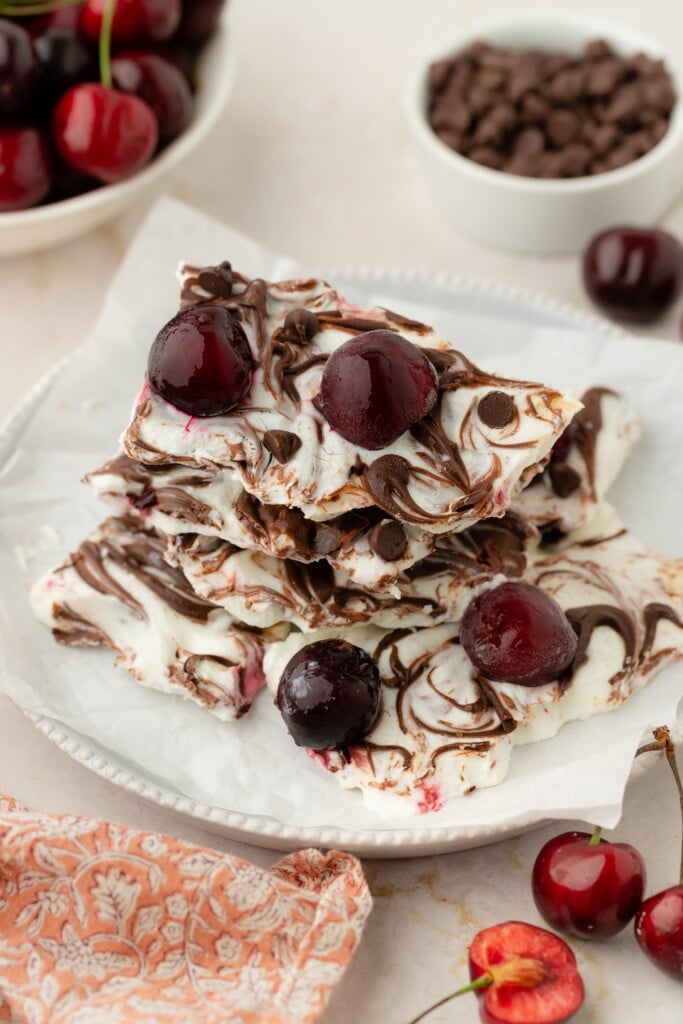 White chocolate bark with dark chocolate swirls and whole cherries on top, reminiscent of Frozen Chocolate Cherry Yogurt Bark, stacked on a plate and surrounded by fresh cherries, chocolate chips, and an orange patterned napkin.